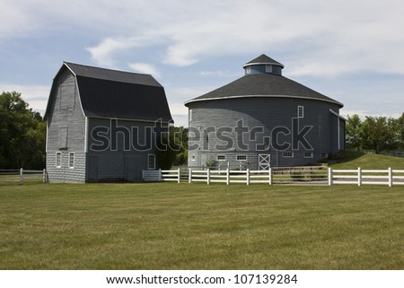 two old barns one round and one traditional with a white fence grass and trees