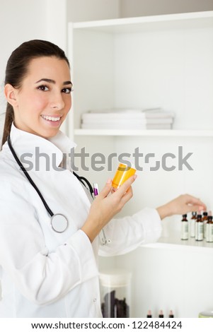 Beautiful young doctor standing by shelf with medicines and holding a orange bottle.
