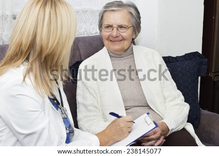 Smiling gray haired  grandmother with crossed fingers sitting on the sofa and talking with her mature female doctor at home.