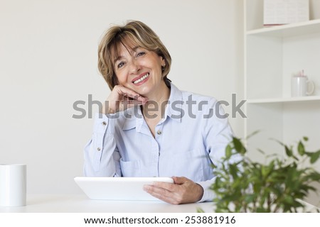 Smiling brown haired woman with hand on her chin sitting at the table in the living room holding  a digital tablet and looking at the camera.