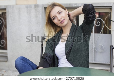 Beautiful blond haired young woman sitting in street cafe with hand behind a head and looking away.