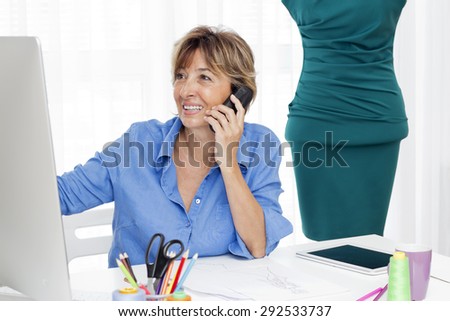 Mature brown haired  fashion woman  designer sitting at the desk talking by phone and looking at a computer monitor.