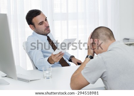 Mid adult manager sitting at the desk opposite his employee and giving him a letter of job termination.