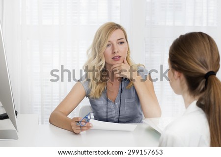 Female   manager with hand on her chin   sitting at the desk opposite young woman and talking.