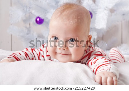 Smiling baby boy on the soft white pillow under the decorated Christmas tree