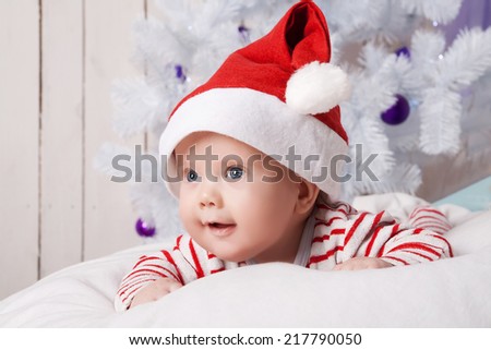 Smiling baby boy in Santa`s hat on the soft white pillow under the decorated Christmas tree