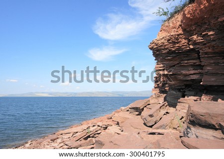 Stone shore of Krasnoyarsk reservoir on the river Yenisei. Sunny day, blue sky, clear water, red stones on the shoreline. Siberian nature landscape, Russia. July 25, 2015.