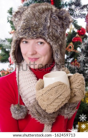 Smiling woman in fluffy hat and mitten under Christmas tree with cup