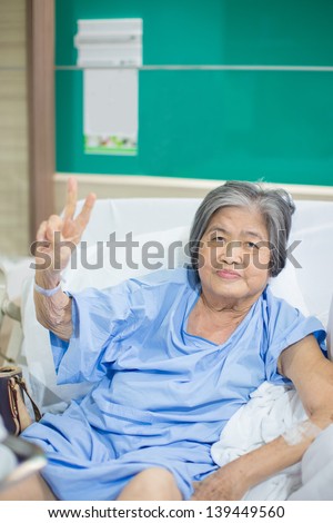 Old cheerful asian granny admitted in hospital holding up 2 fingers sign of courage and will to fight