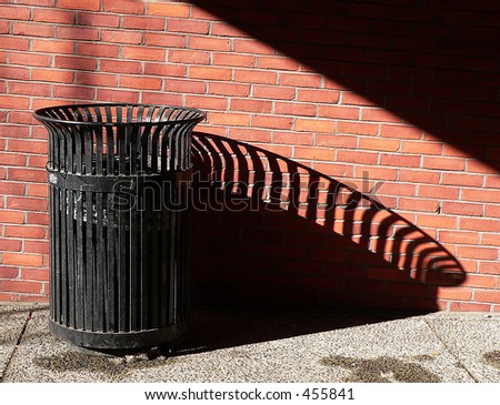 black metal trashcan casts a long shadow, against red brick wall
