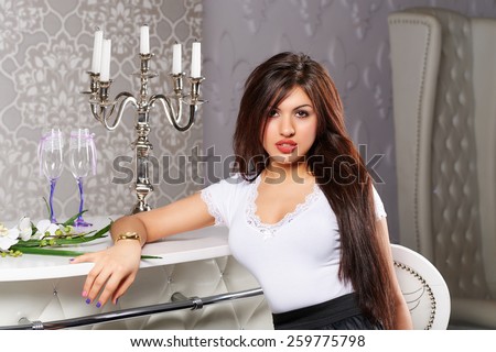 Beautiful leggy brunette girl with big breasts, full lips in a short black dress with a white collar and cuffs in high heels standing near bouquets of roses in vases in white interior