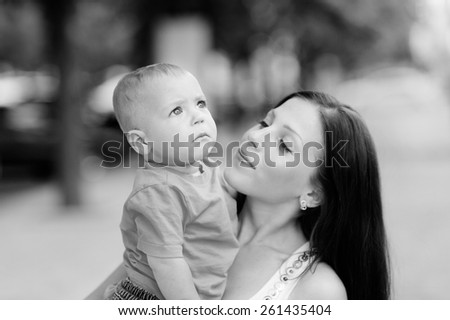 Black and white, 1 year baby in the arms of mother, blond, gray eyes, jeans, Mum young beautiful brunette with straight hair, a white T-shirt. Young mother with her baby in her arms.
