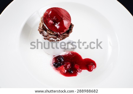 Chocolate cake baked in a mug decorated with chocolate circular plate, painted in red stripes, berry jam, black and red currants sprinkled with powdered sugar on a white plate on a black background