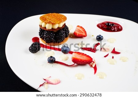 Original unusual berry dessert with honey biscuits, blackberries and butter cream, decorated with strawberries, blueberries, cream, jam with berries, rose petals on a white plate on a black background