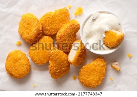 Fried gluten free cornflake crumb chicken nuggets next to a white ceramic bowl of white sauce on white baking paper. Dipped nugget.Top view. Imagine de stoc © 