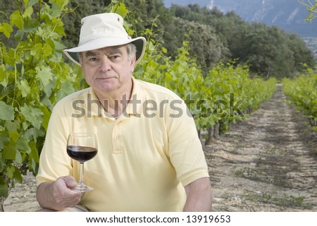 A mature man in a vineyard with a glass of red wine.