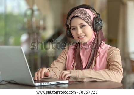 Young pretty Asian muslim woman in head scarf listens to audio with head phone while working on laptop in cafe
