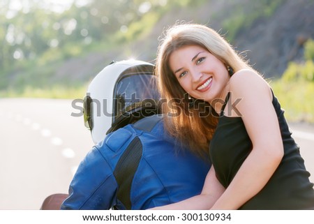 Beautiful young blonde girl in black t-shirt is sitting behind biker man in blue jacket and helmet. Outdoors portrait in bright sunlight.
