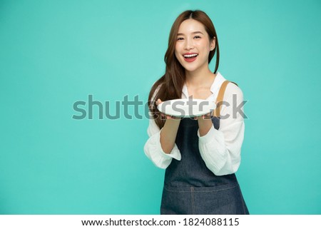 Young Asian woman in apron standing and holding empty white plate or dish isolated on green background