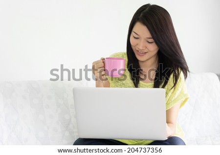 Happy young asian woman using laptop and holding pink cup of coffee indoor.