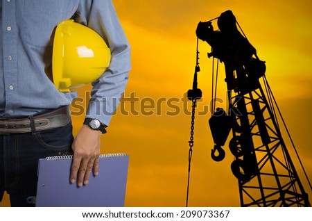 Engineer holding yellow helmet for workers security on background of new highrise apartment buildings and construction cranes on background of evening sunset cloudy sky Silhouette Crane lifts load.