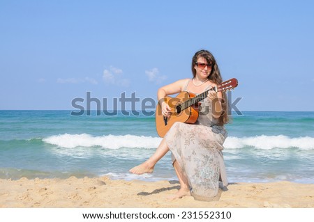 Beautiful long-haired lady in elegant gray dress  playing the guitar on the beach, with sea and blue sky as background