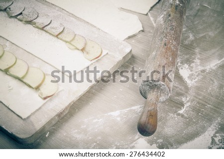 Apple slices on stripes of dough and rolling pin on a light wooden table with flour. Toned.
