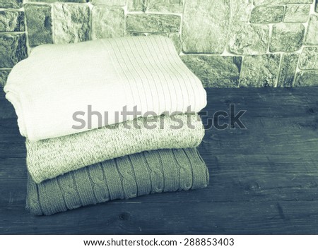 Stack of knitting clothes on wooden table opposite a stony wall. Toned.