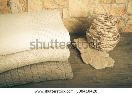 Stack of knitting clothes, hat and mittens on wooden table opposite a stony wall. Toned.