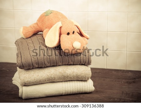 Knitted toy dog on the stack of knitted clothes on wooden table opposite a light tile wall. Toned.
