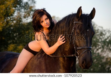 A beautiful young girl horse rider modeling underwear on top of a big and stocky Frisian horse with the sun setting.