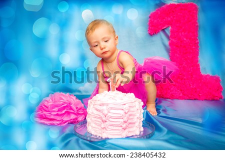 Adorable cute laughing blond hair baby girl with flower head band in pink tutu grabbing vanilla sponge cake with pink and purple heart icing while sitting on blue background