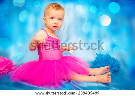 Adorable cute laughing blond hair baby girl with flower head band in pink tutu grabbing vanilla sponge cake with pink and purple heart icing while sitting on blue background