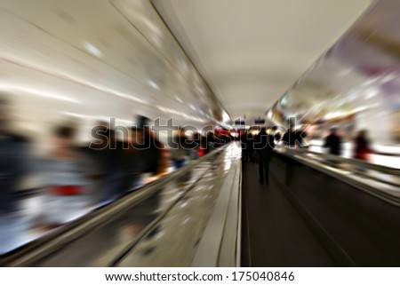 The railing of an escalator in a subway tunnel with passing people photographed with long-term exposure / Subway tunnel