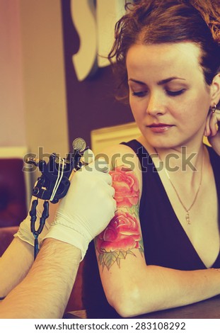 tattooer showing process of making a tattoo on young beautiful hipster woman with red curly hair arm. Tattoo design in the form of rose. Vintage picture