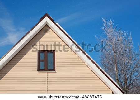 Snow roof of a cottage with a window and tree on a background of the sky with clouds. A falling snow.