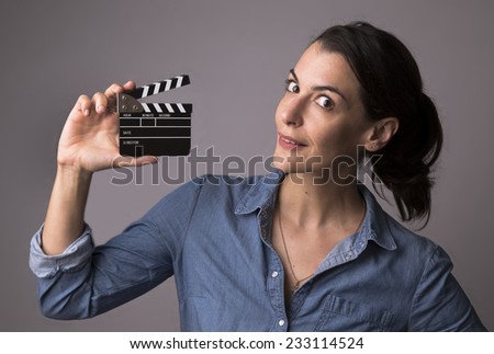 Smiling attractive woman in jeans shirt holding a movie clapper with gray studio background