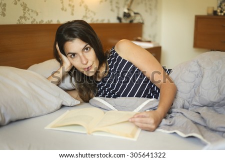 Attractive woman with long brunette hair lying reading a book in bed by the light of an overhead lamp as she snuggles under a blanket
