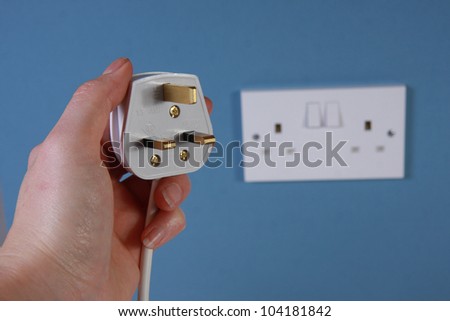 Hand holding a 3 pin plug with socket in the background on a blue wall.  This is a United Kingdom 3 pin plug