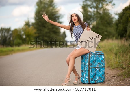sexy brunette woman in shorts , t-shirt and white hat with cardboard sitting on bag hitch hiking on road