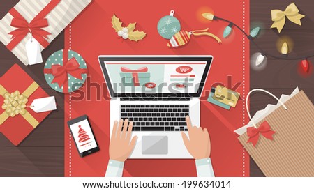 Man purchasing Christmas gifts online using a laptop on his desk, shopping bags and decorations all around, holiday and celebrations banner