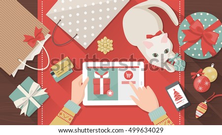 Woman purchasing Christmas gifts online using a tablet, her cat is playing with a bauble on the desk, holiday and celebrations banner