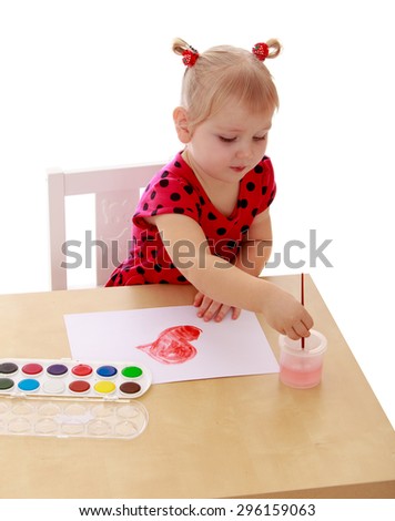 The little blonde girl in a red summer dress polka dot lowers the brush with water, girl paints with watercolors sitting at the table - isolated on white background