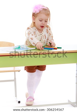 Stylish little blonde girl with a pink bow on his head draws with markers on a piece of paper while standing at the table. The girl is dressed in a white blouse and brown shorts-Isolated on white