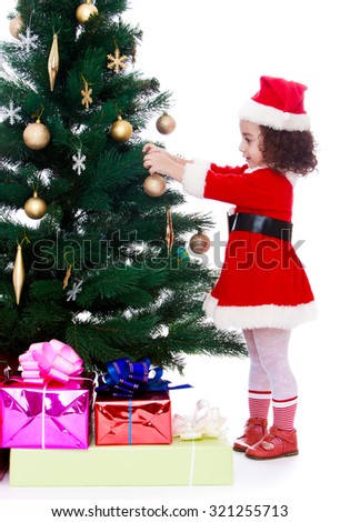 New year mood. Pretty little curly dark haired girl in a suit and cap of Santa Claus hanging toy for Christmas tree. Pared the Christmas tree are boxes with gifts.-Isolated on white background