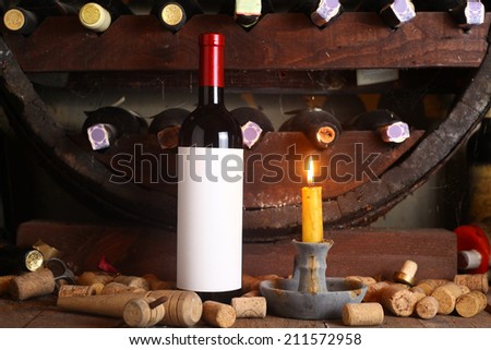 Bottle of red wine with blank label in wine cellar with used corks, bottle shelves and a burning candle