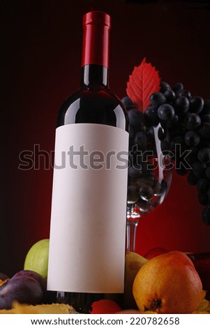 Bottle of red wine with a blank label with a glass filled with ripe grapes standing on autumn leaves with an assortment of ripe fruits over a dark red background