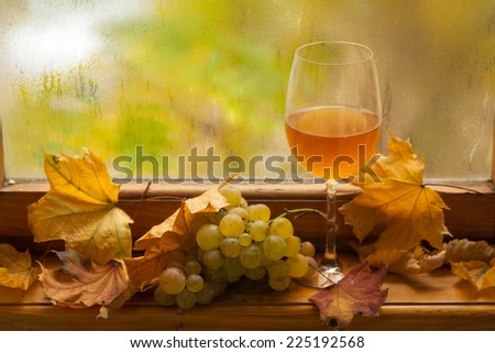 Glass of white wine standing on windowsill with autumn leaves and white grapes with a fogged window in the background