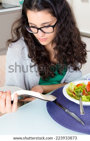 Pretty girl with horn rimmed glasses reading a book while eating salad in the kitchen