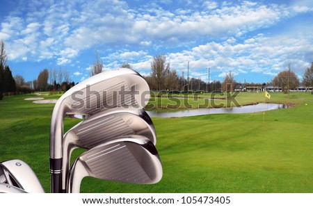 New golf clubs (irons) studio shot with city golf course and driving range as background. Bunkers, green, flag and lake visible.